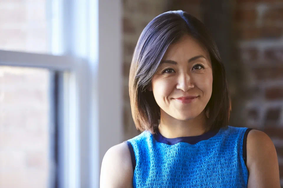 A smiling woman in a blue top, standing by a bright office window
